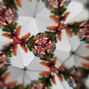May include: Abstract image of a kaleidoscope's interior. The design features a symmetrical pattern with orange, green, and red elements against a white background. Small, reflective silver pieces are also visible, creating a complex and colorful visual.
