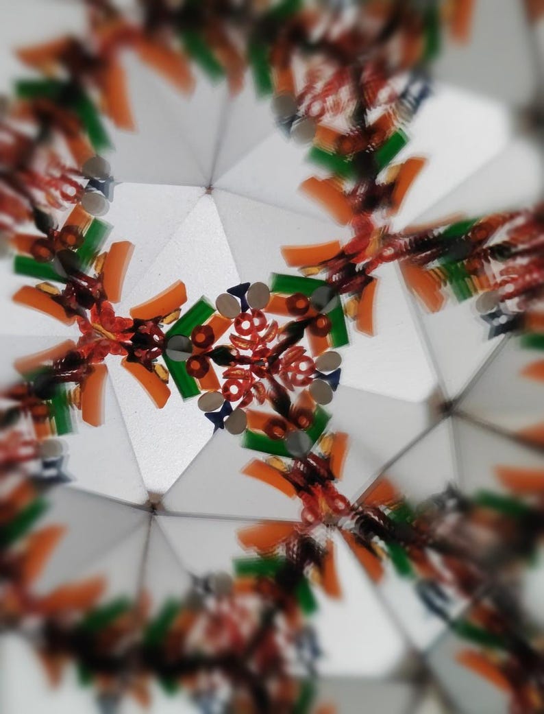 May include: Abstract image of a kaleidoscope's interior. The design features a symmetrical pattern with orange, green, and red elements against a white background. Small, reflective silver pieces are also visible, creating a complex and colorful visual.