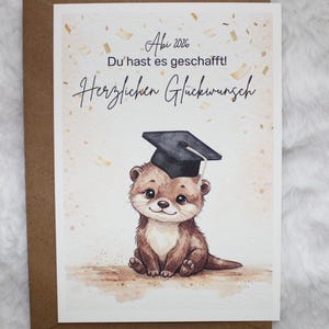 Könnte beinhalten: Eine Abschluss-Glückwunschkarte mit einer Aquarell-Illustration eines Otters mit Doktorhut. Der Text in deutscher Sprache lautet: "Abi 2026, Du hast es geschafft! Herzlichen Glückwunsch."