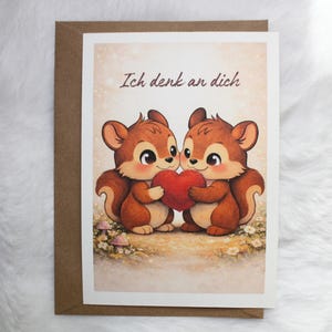 Könnte beinhalten: Eine Grußkarte mit zwei braunen Eichhörnchen, die gemeinsam ein rotes Herz halten. Der Text Ich denk an dich steht über den Eichhörnchen. Der Hintergrund zeigt eine helle Wiese mit kleinen Blumen und Pilzen im unteren Bereich.