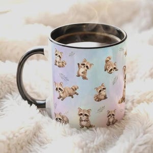 Tasse mit schwarzem Henkel und schwarzer Innenseiten. Bedruckt mit pastellfareben lila, rosa und blau Tönen. Darauf mehrere süße Comic Waschbären in unterschiedlichen Posen.