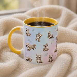 May include: A ceramic mug with a bright yellow handle and rim, featuring a repeating pattern of small, illustrated raccoons in various poses against a soft, pastel-colored background. The mug contains a dark liquid with visible rising steam.
