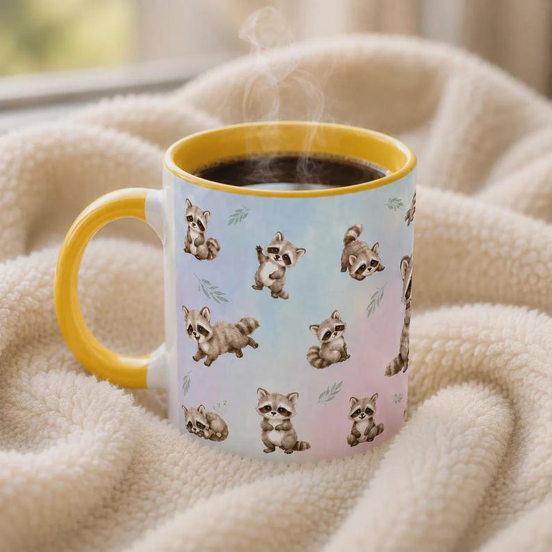 May include: A ceramic mug with a bright yellow handle and rim, featuring a repeating pattern of small, illustrated raccoons in various poses against a soft, pastel-colored background. The mug contains a dark liquid with visible rising steam.