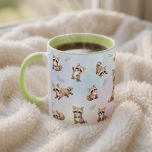 May include: A ceramic mug with a light green handle and rim, featuring a repeating pattern of small, illustrated raccoons in various poses against a soft, pastel-colored background. The mug is filled with a dark brown liquid, emitting a faint steam.