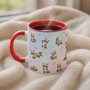 May include: A ceramic mug with a bright red handle and interior rim, featuring a repeating pattern of small, illustrated brown and grey raccoons in various poses against a soft, light blue and pink gradient background with green leaf accents.
