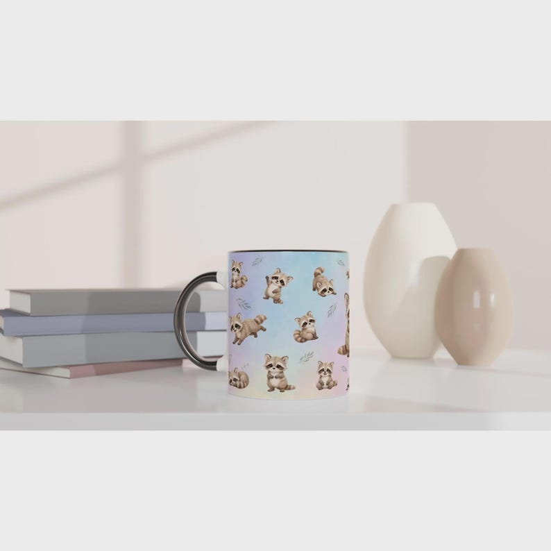 May include: A ceramic mug with a black handle and rim, featuring a repeating pattern of small, illustrated brown and grey raccoons in various poses against a soft, pastel-colored gradient background of light blue, pink, and lavender hues.