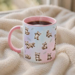 May include: A ceramic mug with a light pink handle and rim, featuring a repeating pattern of small, illustrated raccoons in various poses against a soft, pastel-colored background. The mug is filled with a dark liquid, and steam rises from the top.