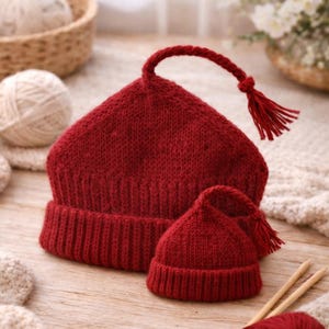 May include: Two knitted red hats, one adult-sized and one smaller, are displayed on a wooden surface. The larger hat features a long, twisted cord with a tassel. A ball of red yarn and knitting needles are nearby, indicating a handmade item.