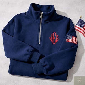 May include: Navy blue quarter-zip sweatshirt with a red monogram and an American flag patch on the sleeve. The sweatshirt has a high collar and ribbed cuffs. A small American flag is also visible.