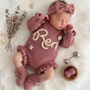 May include: A dusty rose knitted romper featuring the name Ren in white cursive yarn lettering, accented with small white flowers. The set includes a matching headband with a large bow, knitted booties with tie closures, and a small ball of yarn.
