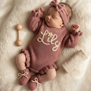May include: A dusty rose knitted baby romper featuring ruffled edges, long sleeves, and the name Lily embroidered in white yarn with a small flower accent. The set includes a matching knitted headband with a bow and a pair of knitted booties.