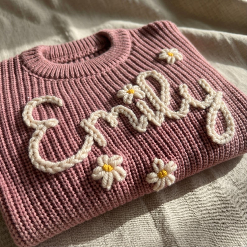 May include: A dusty rose-colored knit sweater with the name "Emily" embroidered in cream-colored yarn. The sweater is decorated with small, cream-colored daisy appliques with yellow centers. The sweater is folded on a neutral-colored surface.