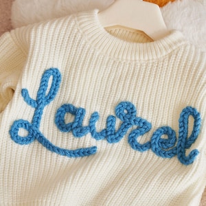 May include: Cream-colored knit sweater with the word "Laurel" in blue, textured, cursive lettering. The sweater is hanging on a white hanger. The knitwear has a ribbed texture.