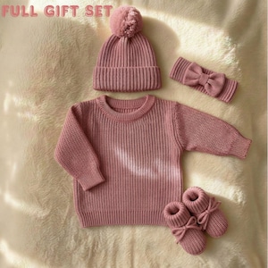 May include: A full gift set of knitted baby clothing in a dusty rose color. The set includes a beanie with a pom-pom, a sweater, a headband with a bow, and booties. The items are arranged on a soft, textured surface.