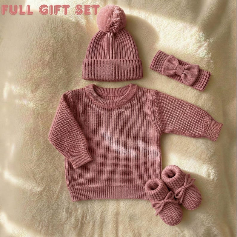 May include: A full gift set of knitted baby clothing in a dusty rose color. The set includes a beanie with a pom-pom, a sweater, a headband with a bow, and booties. The items are arranged on a soft, textured surface.