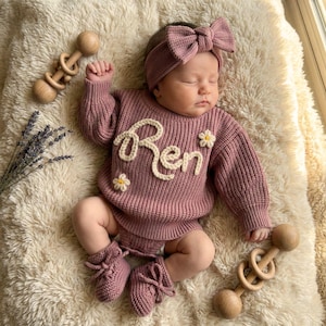 May include: A sleeping baby wearing a mauve knit sweater and shorts set with the name "Ren" in cream-colored yarn. The outfit includes a matching headband, booties, and small daisy accents. Wooden baby rattles are placed nearby.