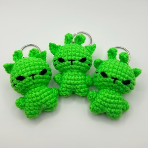 May include: Three handmade green crochet alien keychains. Each keychain features a small, green alien with black eyes and a T-shaped mouth. The aliens have small arms and legs, and are attached to silver key rings.