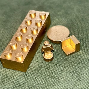 Puede incluir: Una colección de ladrillos Lego y una minifigura Lego de color dorado. El conjunto incluye un ladrillo rectangular, un disco redondo, una forma geométrica y una pequeña figura Lego, todo con un acabado dorado metálico.