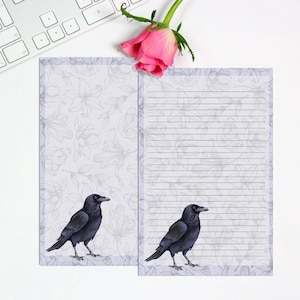 May include: Two notepads with a floral design and a raven illustration. One notepad is lined, and the other is blank. A pink rose and a keyboard are in the background. The notepads have a light blue border.