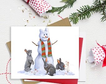 May include: A greeting card featuring a watercolor illustration of a snowman with an orange carrot nose and a red and green striped scarf, standing in snow with three grey rabbits. The text Julie Townsend Studio is printed at the bottom.