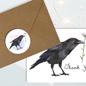 May include: A watercolour illustration of a black crow holding a daisy, with the words "Thank You!" on a white card. A matching sticker and a brown envelope are also present. The crow is the focal point, with detailed feathers and a delicate flower.