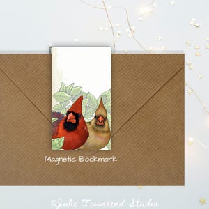 May include: A rectangular magnetic bookmark featuring a detailed illustration of a bright red male cardinal and a tan female cardinal perched among green leaves. The bookmark is positioned on a brown envelope with the text Magnetic Bookmark.
