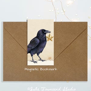 May include: A rectangular magnetic bookmark featuring a detailed illustration of a black raven holding a gold star in its beak. The bookmark is displayed on a brown kraft paper envelope with the text Magnetic Bookmark printed below it.