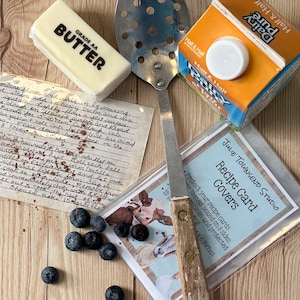 May include: A close-up of a recipe card cover with a cow illustration, a handwritten recipe card, a stick of butter, a metal slotted spoon, and a carton of half and half cream. The recipe card cover is clear plastic and has the text "Recipe Card Covers" and "Julie Townsend Studio".