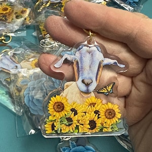 May include: A clear acrylic keychain featuring a goat with a blue and white face, a yellow butterfly, and sunflowers. The keychain has a gold-colored ring. Other keychains with similar designs are visible in the background.
