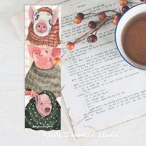 May include: A bookmark featuring three illustrated pigs in cozy attire. The top pig wears a plaid scarf, the middle pig a plaid shawl, and the bottom pig a green polka-dot hooded cloak. A cup of coffee and a book are in the background.