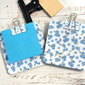May include: Two memo clipboards with a white background and a blue floral pattern. One clipboard holds a blue sticky note. A black binder clip and a small, square, tan sticky note are also visible.