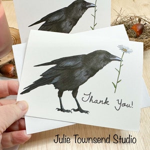May include: White greeting cards with a detailed illustration of a black crow holding a daisy. The words "Thank You!" are handwritten on the card. The artist's name, "Julie Townsend Studio," is printed at the bottom.