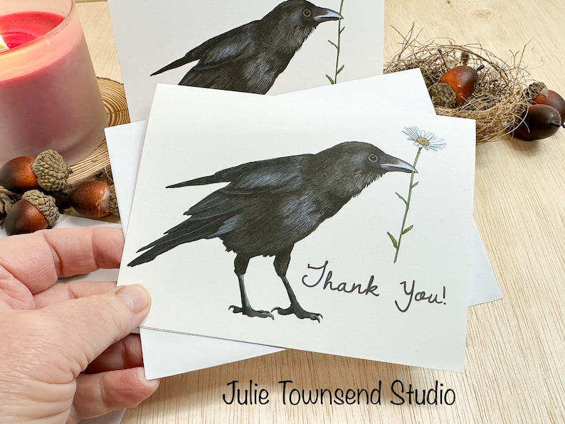May include: White greeting cards with a detailed illustration of a black crow holding a daisy. The words "Thank You!" are handwritten on the card. The artist's name, "Julie Townsend Studio," is printed at the bottom.