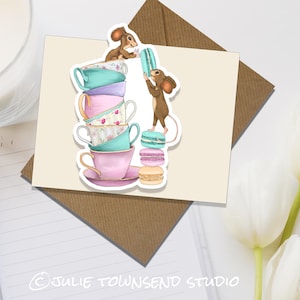 May include: Two mice stacking teacups and macarons. The teacups are decorated with floral patterns and are stacked in a tower. The macarons are pastel pink and green. The mice are brown and have white accents.
