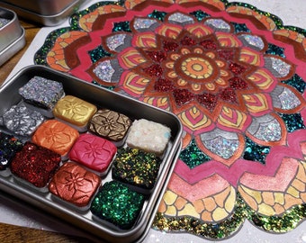 May include: A metal tin with 12 small squares filled with different colors of glitter and metallic paint. The tin is open and the squares are visible. The tin is sitting on a wooden surface next to a large, colorful mandala design.