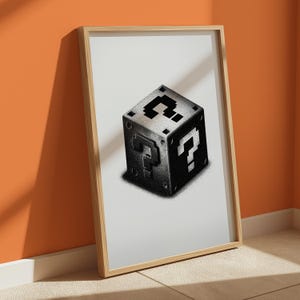 May include: A framed print depicting a pixelated question mark cube, a design from a well-known video game. The cube is in shades of grey and black, with a shadow effect. The frame is made of light-coloured wood.