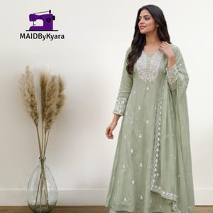 May include: A sage green long-sleeved dress featuring white floral embroidery around the neckline and cuffs, with a matching sheer dupatta draped over the shoulder. The text MAIDByKyara appears in the upper left corner with a sewing machine icon.