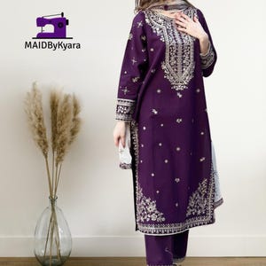 May include: A deep purple embroidered salwar kameez set. The long-sleeved tunic and matching pants feature intricate gold-colored embroidery along the neckline, cuffs, and hem. The set includes a matching scarf. The logo "MAIDByKyara" is in the top left corner.