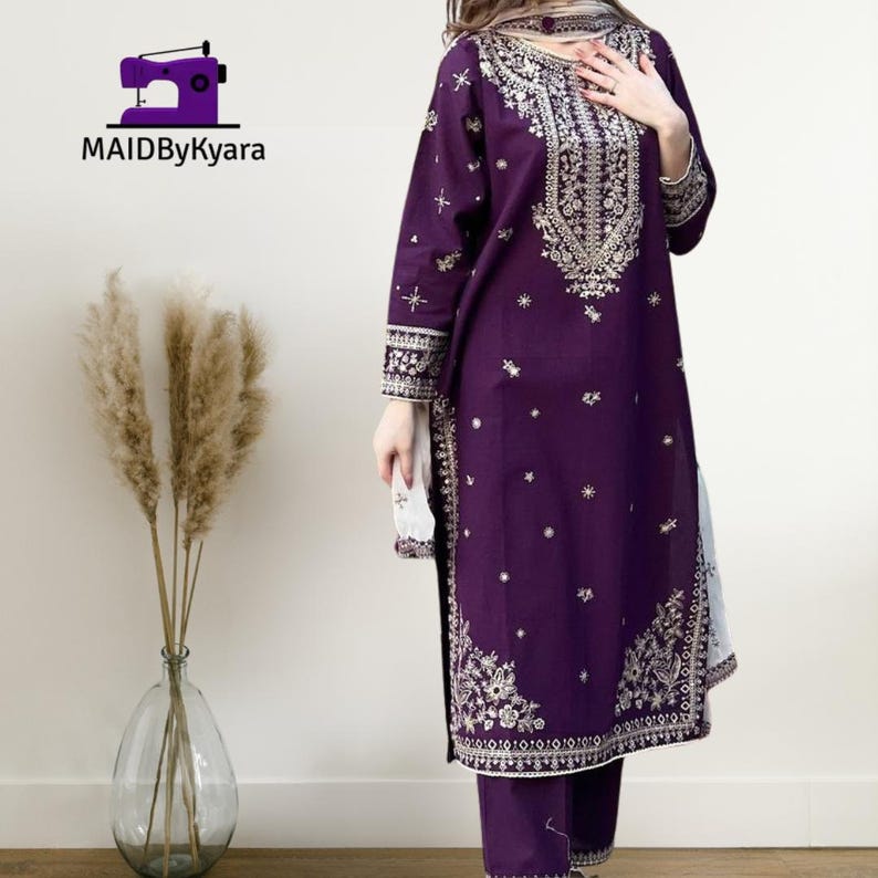 May include: A deep purple embroidered salwar kameez set. The long-sleeved tunic and matching pants feature intricate gold-colored embroidery along the neckline, cuffs, and hem. The set includes a matching scarf. The logo "MAIDByKyara" is in the top left corner.