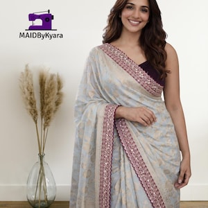 Puede incluir: Un sari azul claro con un estampado floral y un borde burdeos adornado con pequeños adornos blancos. El sari está drapeado sobre un top morado oscuro. La imagen incluye el texto "MAIDByKyara" y un logotipo de máquina de coser.