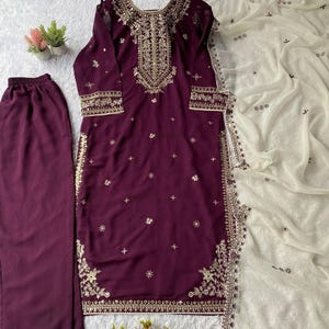 May include: A plum-colored embroidered salwar kameez set with matching pants and a white dupatta. The tunic and pants feature gold embroidery along the neckline, cuffs, and hem. The dupatta has delicate embroidery.