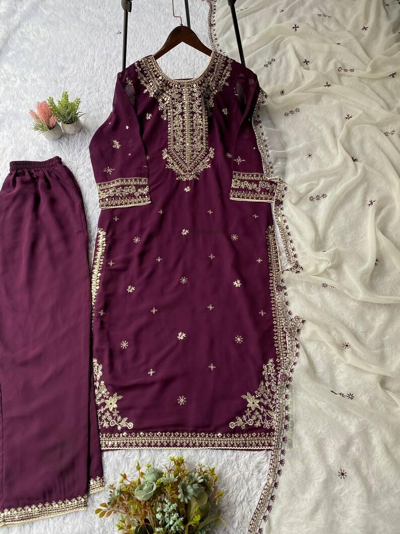 May include: A plum-colored embroidered salwar kameez set with matching pants and a white dupatta. The tunic and pants feature gold embroidery along the neckline, cuffs, and hem. The dupatta has delicate embroidery.