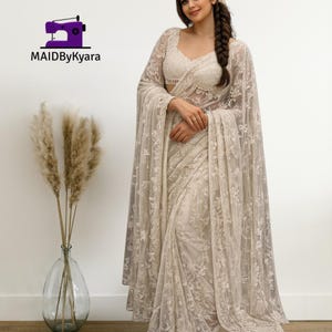 Puede incluir: Un sari bordado de color crema con una blusa y una capa a juego. El sari presenta intrincados patrones de encaje floral. La imagen incluye el texto "MAIDByKyara" y un logotipo de máquina de coser.