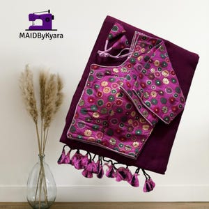 Può includere: Un sari color prugna con una camicetta abbinata e un piccolo accessorio. La camicetta presenta un motivo floreale nei toni del rosa, verde e oro. Il sari ha un bordo con nappe. Il logo "MAIDByKyara" si trova in alto a sinistra.