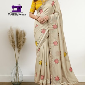 Może przedstawiać: Beżowe sari z różowymi i żółtymi haftami kwiatowymi. Sari ma ozdobną bordiurę i jest połączone z żółtą bluzką. Logo "MAIDByKyara" jest widoczne w lewym górnym rogu.