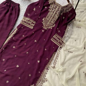 May include: A plum-colored, embroidered salwar kameez set. The long-sleeved tunic and matching pants are adorned with gold embroidery and small, scattered embellishments. A cream-colored, sheer dupatta with embroidered details completes the ensemble.