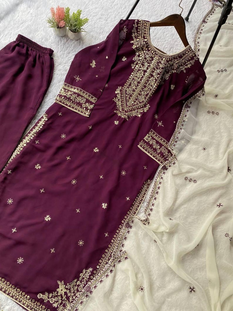 May include: A plum-colored, embroidered salwar kameez set. The long-sleeved tunic and matching pants are adorned with gold embroidery and small, scattered embellishments. A cream-colored, sheer dupatta with embroidered details completes the ensemble.
