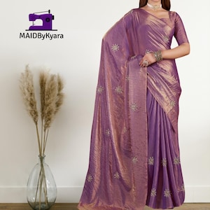 Peut inclure: Un sari violet chatoyant avec des accents dorés et des détails floraux brodés. Le tissu a un aspect texturé, et le design comprend un chemisier assorti. Le logo de la marque "MAIDByKyara" est visible.