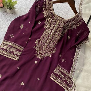 May include: A plum-colored tunic with intricate gold embroidery around the neckline, cuffs, and bodice. The long-sleeved garment features delicate floral and geometric patterns. A matching white scarf with gold trim is draped nearby, suggesting a formal or festive attire.
