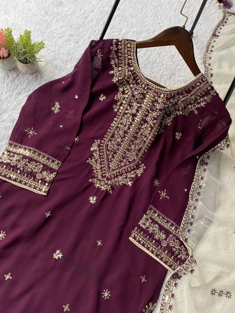 May include: A plum-colored tunic with intricate gold embroidery around the neckline, cuffs, and bodice. The long-sleeved garment features delicate floral and geometric patterns. A matching white scarf with gold trim is draped nearby, suggesting a formal or festive attire.